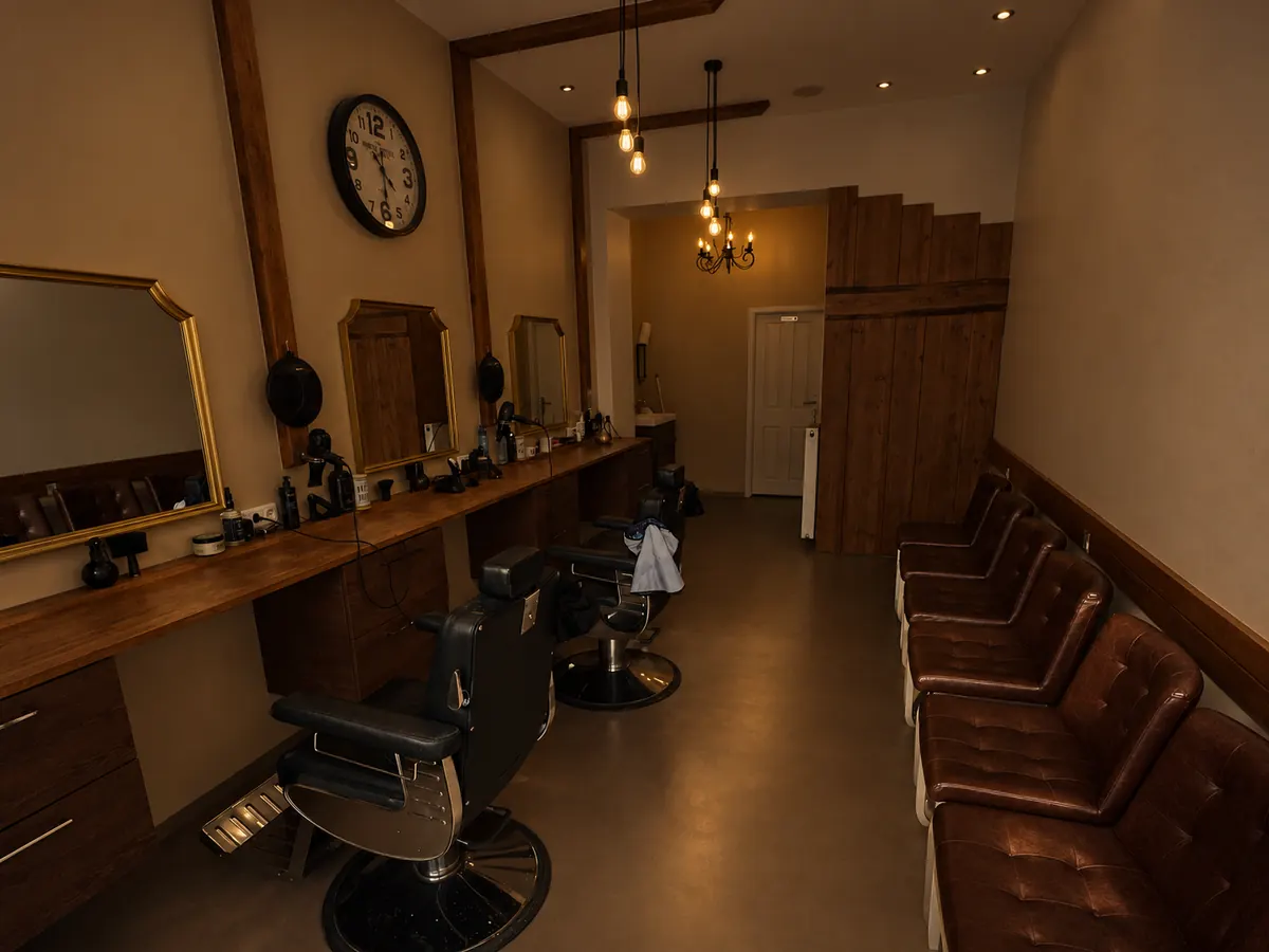 Blick durch den Salon: Barbierstühle an der Spiegelwand, gegenüber braune Wartesessel, hinten eine Holzwand